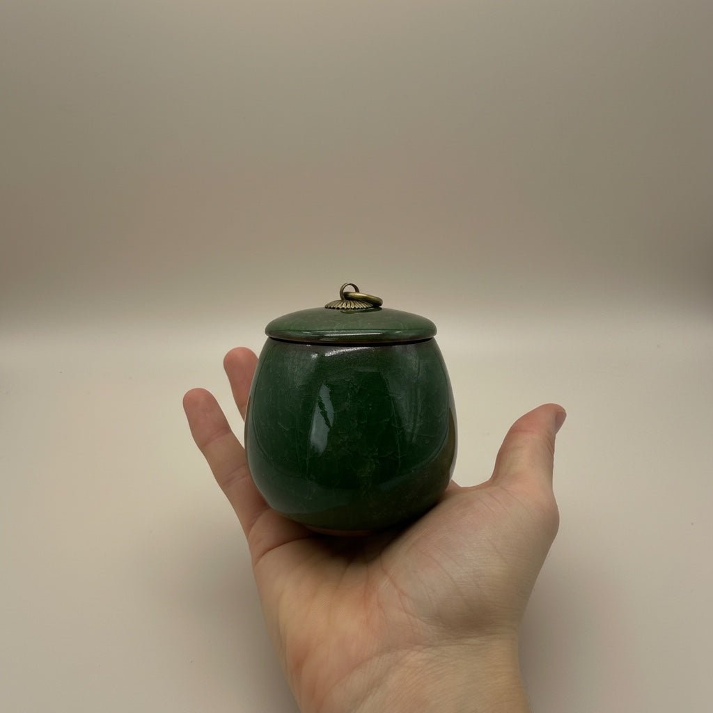 Donkergroene keramische urn met deksel en metalen ring, ovaalvormig, in een hand gehouden