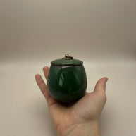 Donkergroene keramische urn met deksel en metalen ring, ovaalvormig, in een hand gehouden