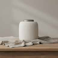 Witte geribbelde keramische urn met een grijze deksel, staand op een houten tafel met linnen doek
