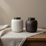 Twee geribbelde keramische urnen, wit en donkerbruin, met donkere deksels op houten tafel