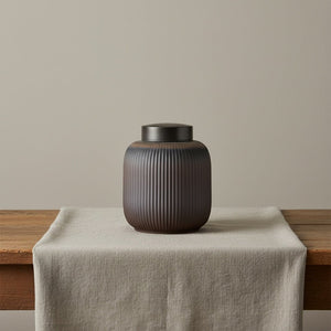 Keramische bruine urn met verticale ribbels, zwarte deksel, op houten tafel met linnen loper