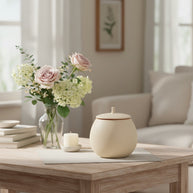 Beige ronde keramische urn met deksel op houten tafel met bloemen en kaars