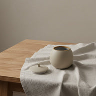 Beige ronde keramische urn met deksel op een lichte linnen doek, houten tafel