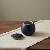 Donkerblauwe, ronde urn met textuur en een beige rand, deksel ernaast op een houten tafel