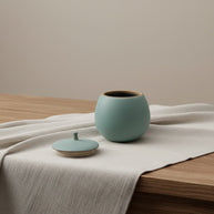 Lichtblauwe, ronde keramische Luna urn met deksel op een houten tafel met linnen doek