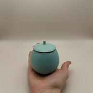 Lichtblauwe ronde urn met deksel en kleine knop, mat oppervlak, in hand gehouden