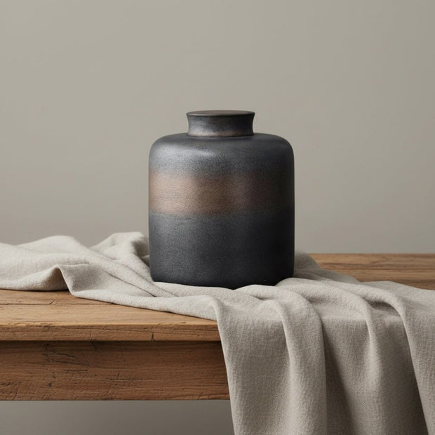 Zwarte keramische urn met koperkleurige band, ronde vorm, op houten tafel met linnen doek