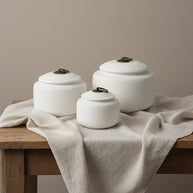 Drie witte, geribbelde, ronde urnen met bronzen handvatten op een houten tafel met linnen doek