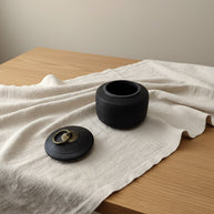 Zwarte geribbelde urn met deksel en gouden handvat, op een houten tafel met lichte stof