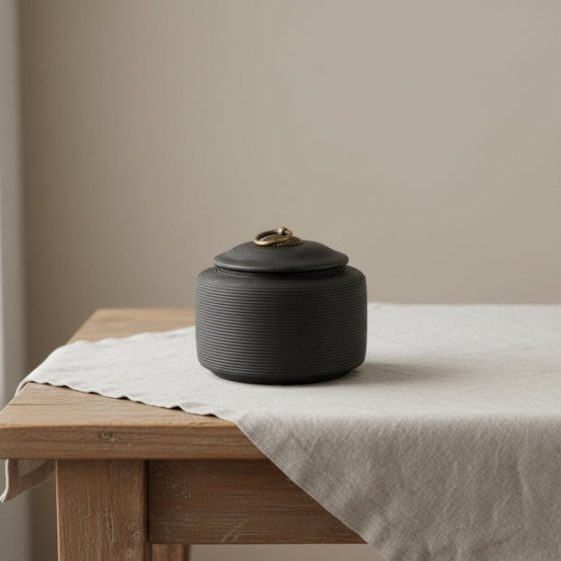 Zwarte keramische urn met horizontale ribbels en een bronzen ring op het deksel