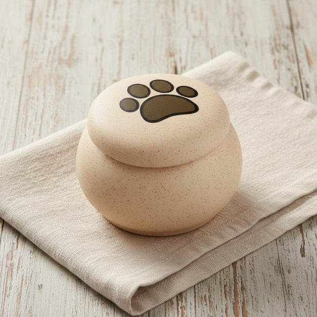 Ronde beige urn met spikkels en een zwarte pootafdruk op de deksel, op een linnen doek