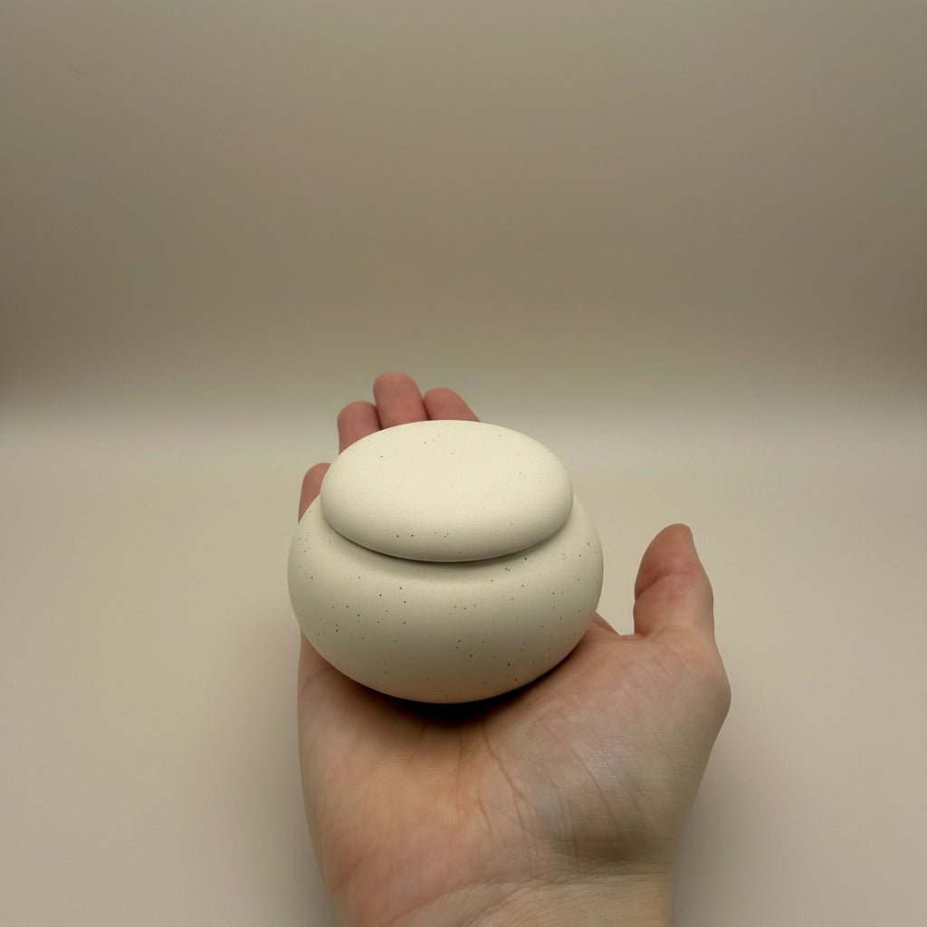 Witte, ronde keramische urn met spikkels, deksel, in de hand gehouden