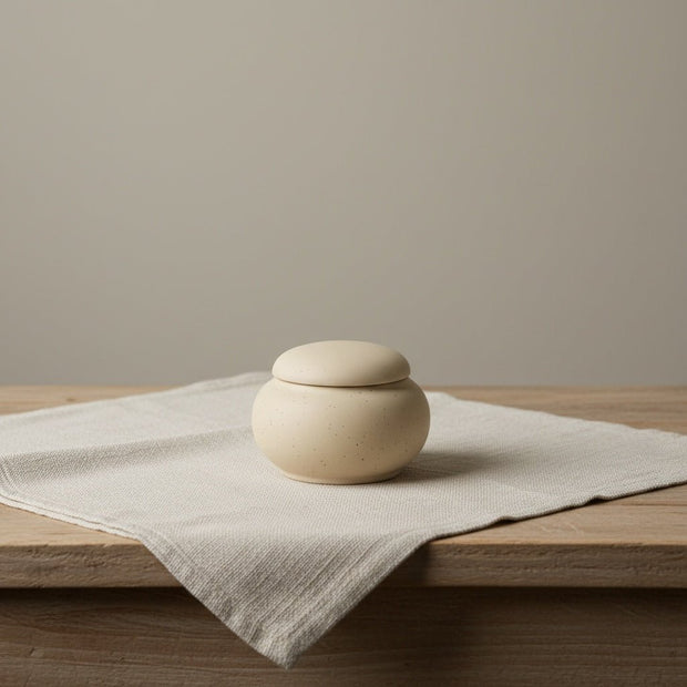 Serena urn, beige gespikkelde keramische urn met ronde vorm en deksel op linnen doek