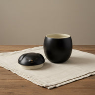Zwarte keramische Shiba urn met gouden details, ronde vorm, op een houten tafel met linnen doek