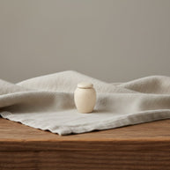 Suna urn, beige keramiek met spikkels, ronde vorm, op lichte stof en houten tafel